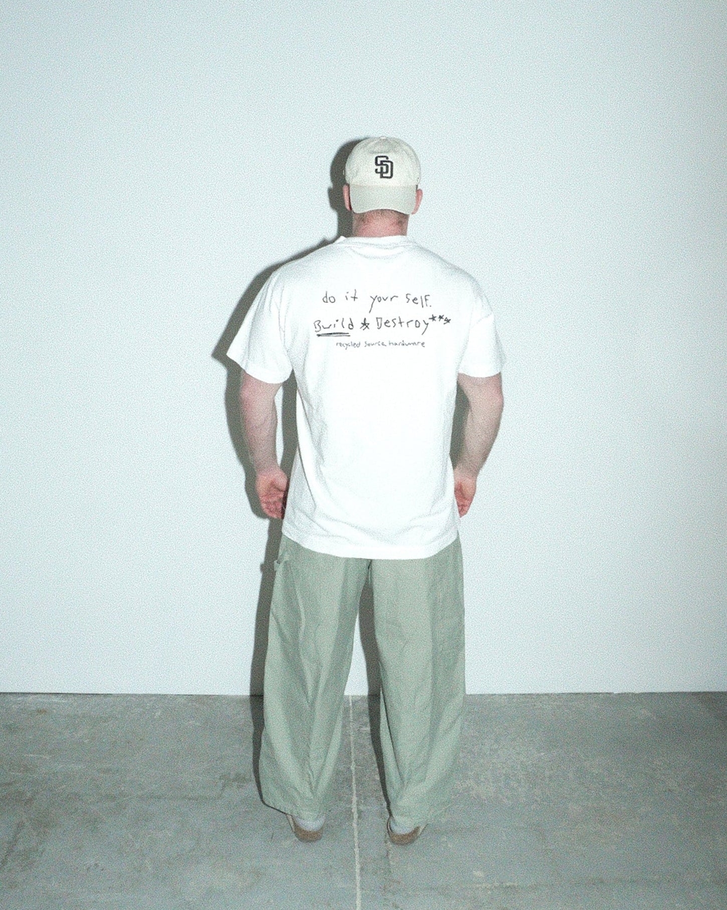 Person wearing a white t-shirt with text and a cap, standing against a plain white wall.